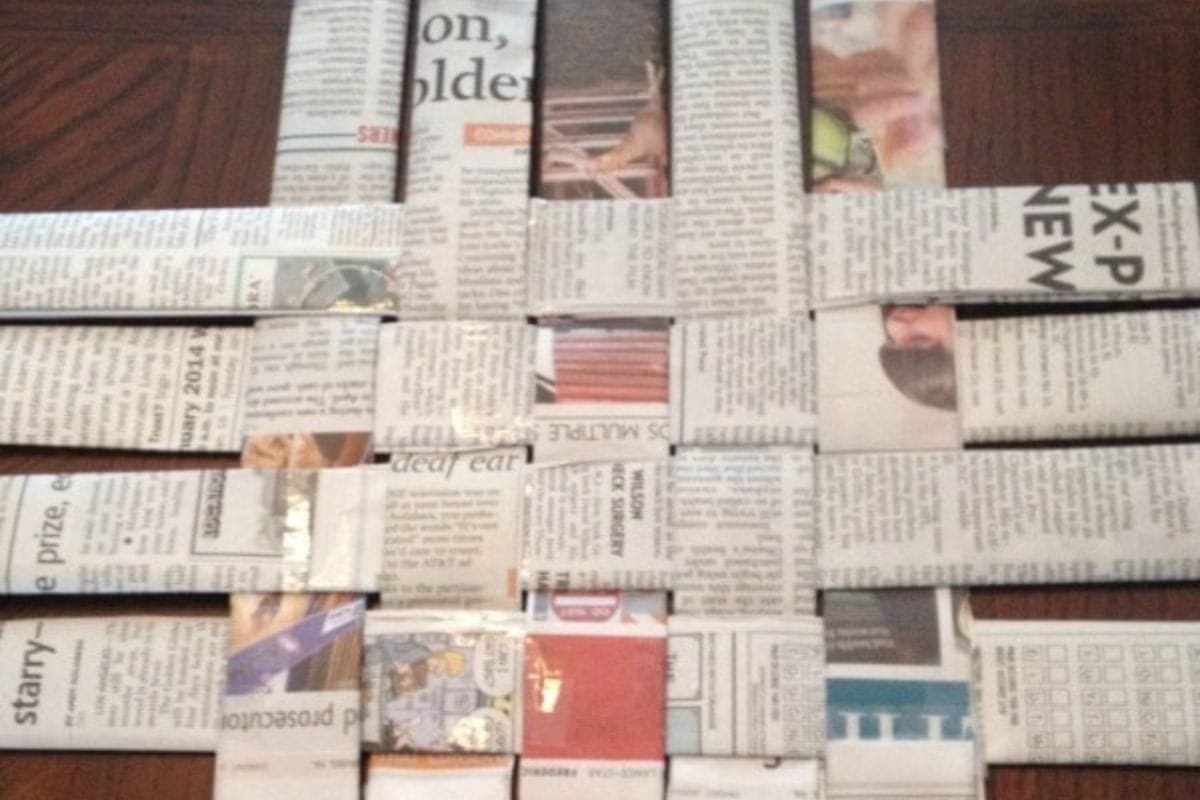 Strips of newspaper are woven together in a grid pattern on a wooden surface, forming the base of a handcrafted newspaper basket.