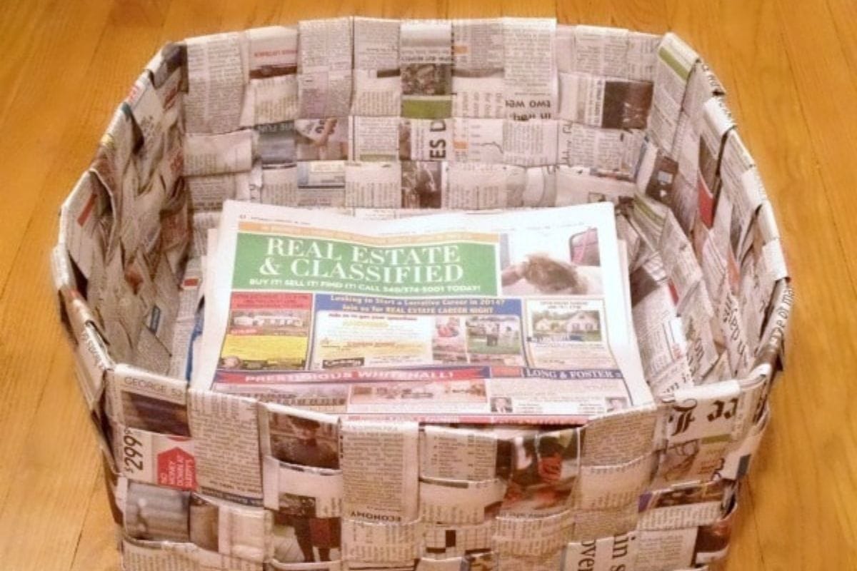 A rectangular newspaper basket made from woven strips sits on a wooden floor, filled with additional newspaper inside the basket.
