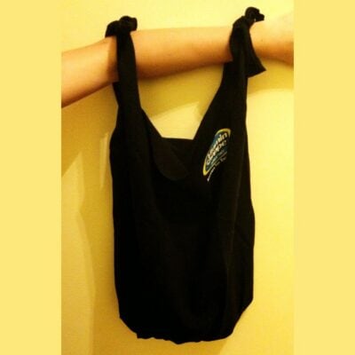 A black t-shirt bag with two knotted handles hangs from a person's arm against a yellow wall. A small, colorful logo stands out on the front of the bag.