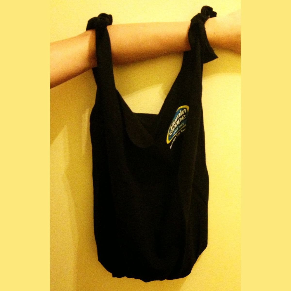 A black t-shirt bag with two knotted handles hangs from a person's arm against a yellow wall. A small, colorful logo stands out on the front of the bag.