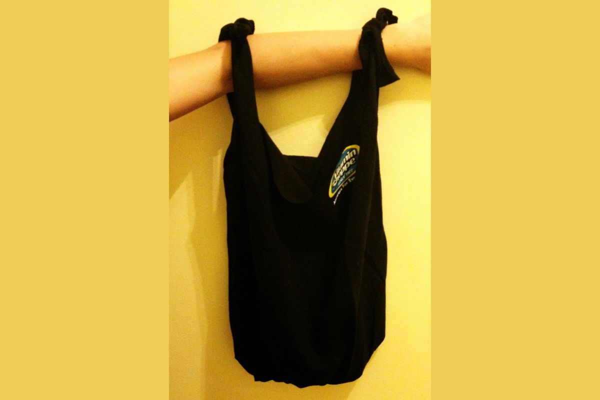 A person's arm holds a black t-shirt bag with knotted handles and a small colorful patch, against a yellow background.