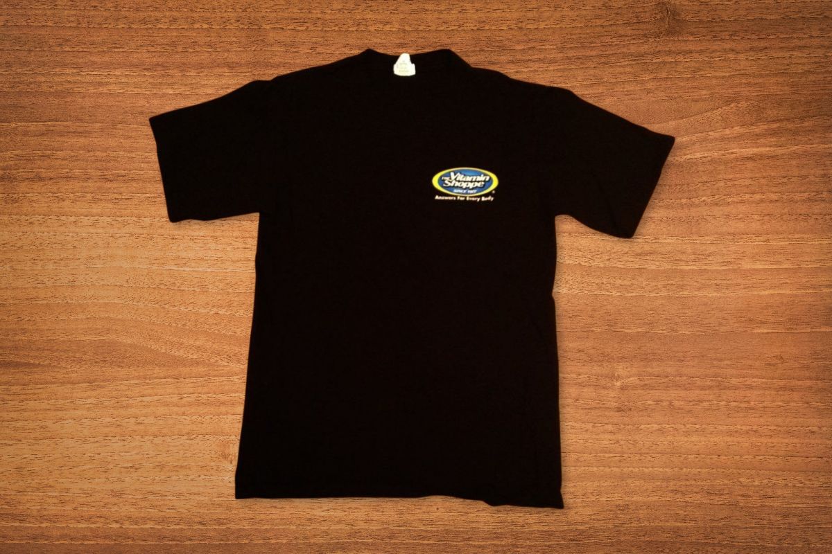 A black short-sleeve t-shirt with a small, oval-shaped logo on the left chest is laid flat on a wooden surface.