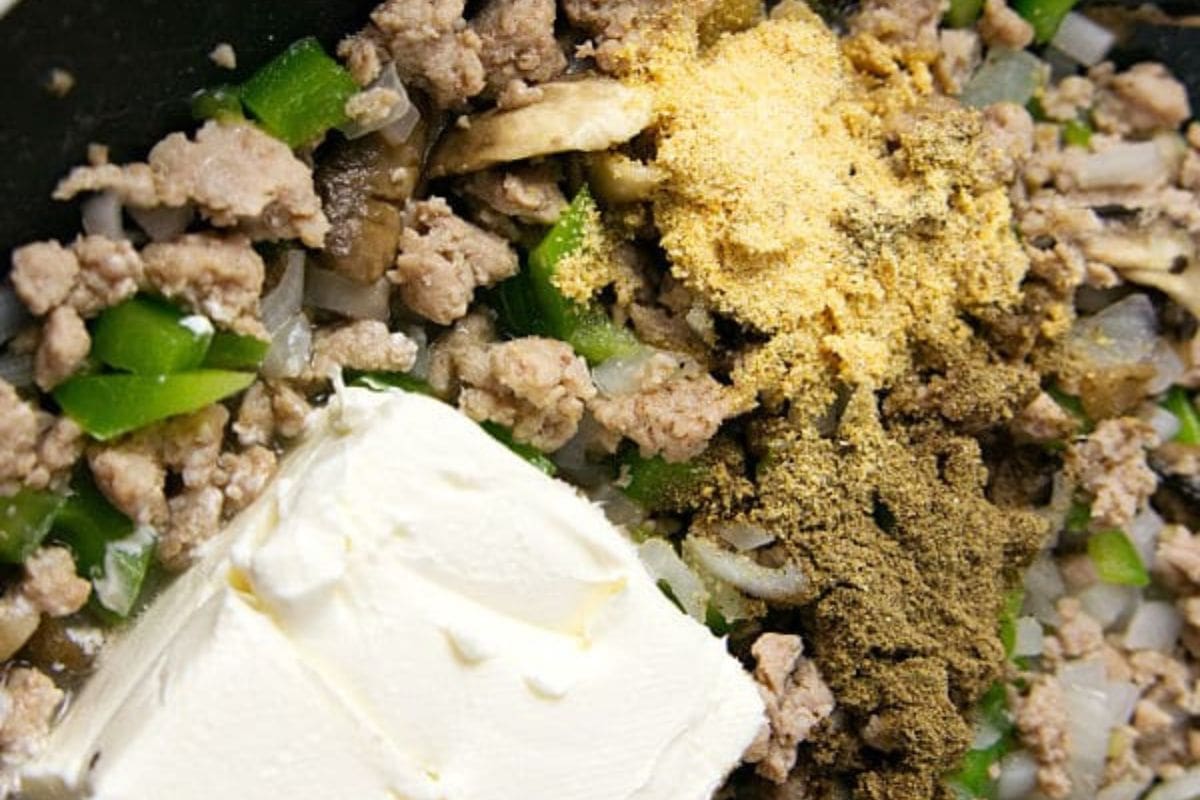 Cream cheese, seasoning, diced bell pepper, mushrooms, onions, and ground turkey cooking in a skillet.