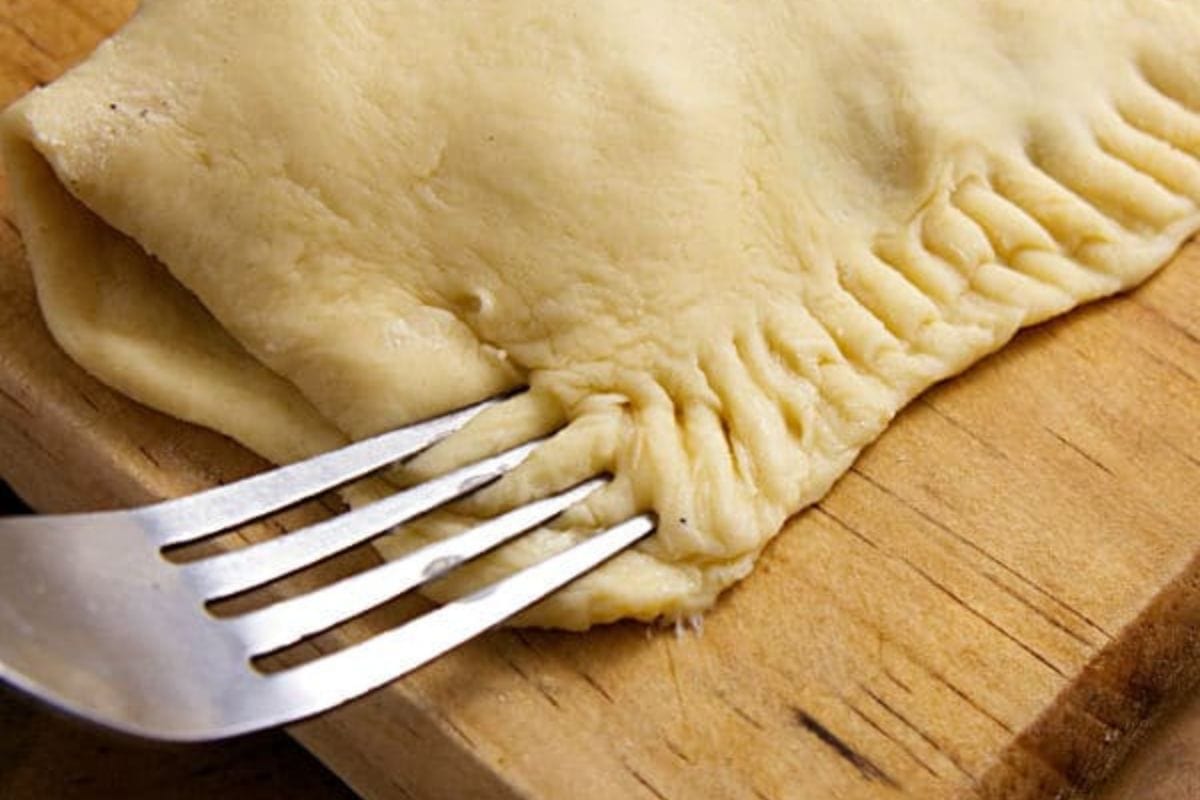 A fork pressing down the edges of the dough of the turkey turnover on a wooden cutting board.