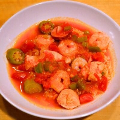 A bowl of healthy gumbo with shrimp, chicken, okra, bell peppers, tomatoes, and rice in a red broth, served in a white dish on a wooden surface.