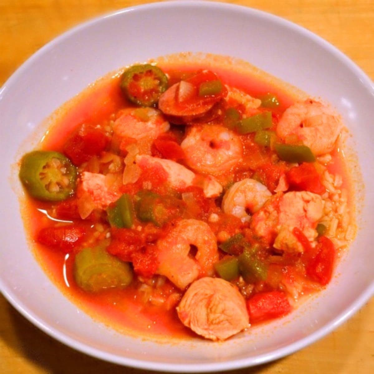 A bowl of healthy gumbo with shrimp, chicken, okra, bell peppers, tomatoes, and rice in a red broth, served in a white dish on a wooden surface.