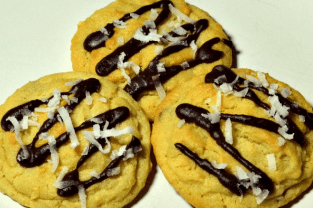 Three Almond Joy cookies topped with drizzled chocolate and shredded coconut rest on a white surface.
