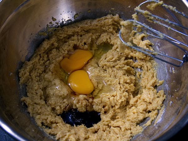 eggs and vanilla added to the cookie batter in a metal mixing bowl with whisks above it