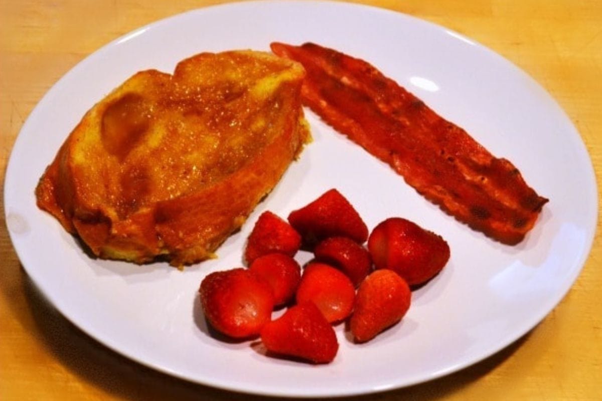 A plate with a slice of baked French toast, a strip of bacon, and several whole strawberries sits on a wooden surface.