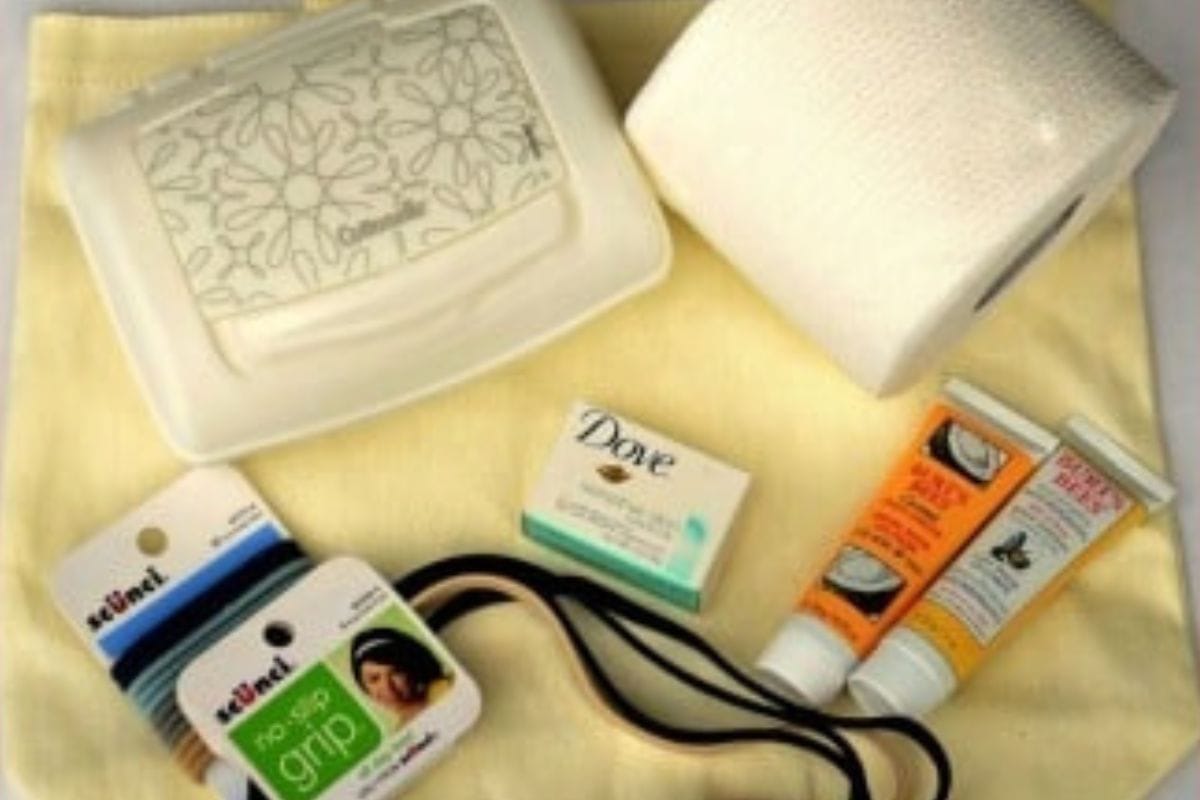 A rolled toilet paper, a pack of wipes, a bar of Dove soap, hair ties, and two tubes of lotion on a folded yellow towel.