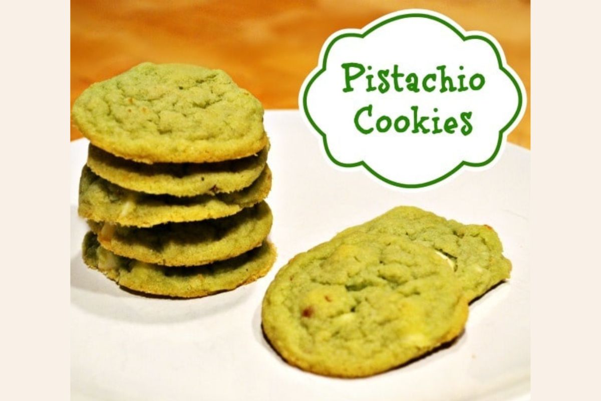 A stack of green pistachio cookies sits on a white plate, with two pistachio cookies lying flat beside the stack. A sign in the background reads "Pistachio Cookies.