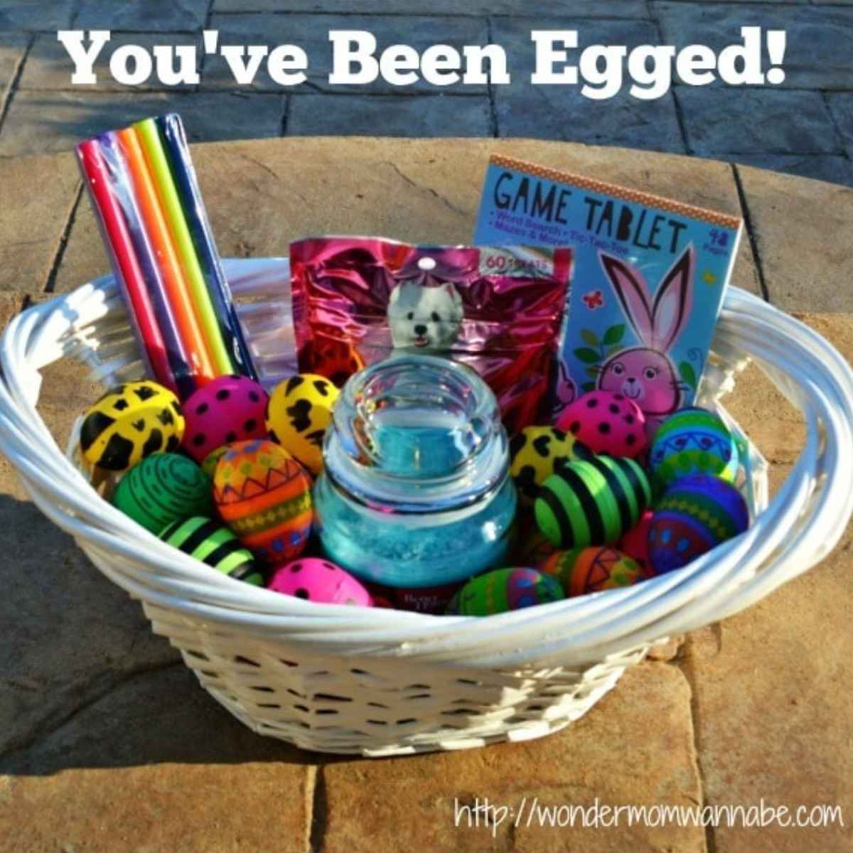 A white basket filled with colorful decorated eggs, candles, a game tablet, candy, and a jar sits below the cheerful text "You've Been Egged!"—perfect for surprising friends or family with an egg-citing "you've been egged" gift.