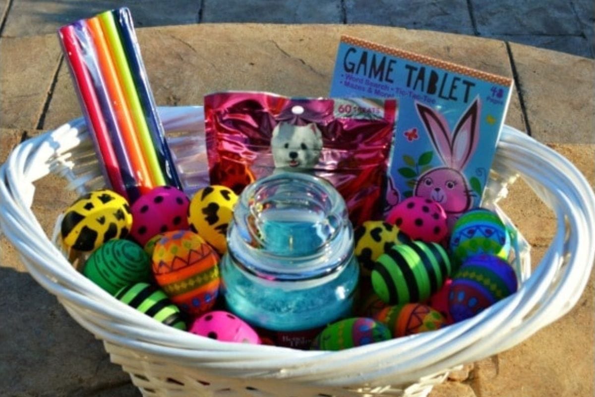 A white basket filled with decorated eggs, a jar, straws, a snack pouch, and a game tablet pack.