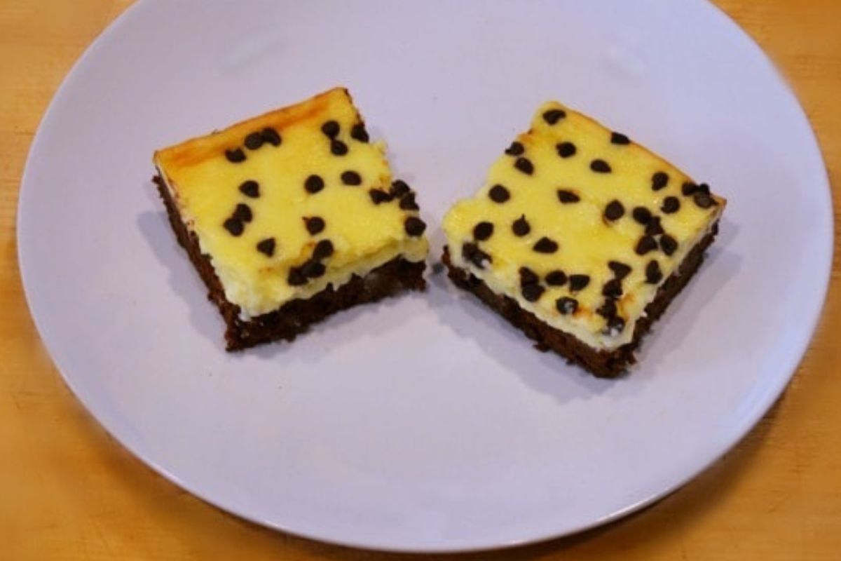Two square guilt-free cheesecake brownies with a yellow cream cheese layer and chocolate chips on top, served on a white plate.