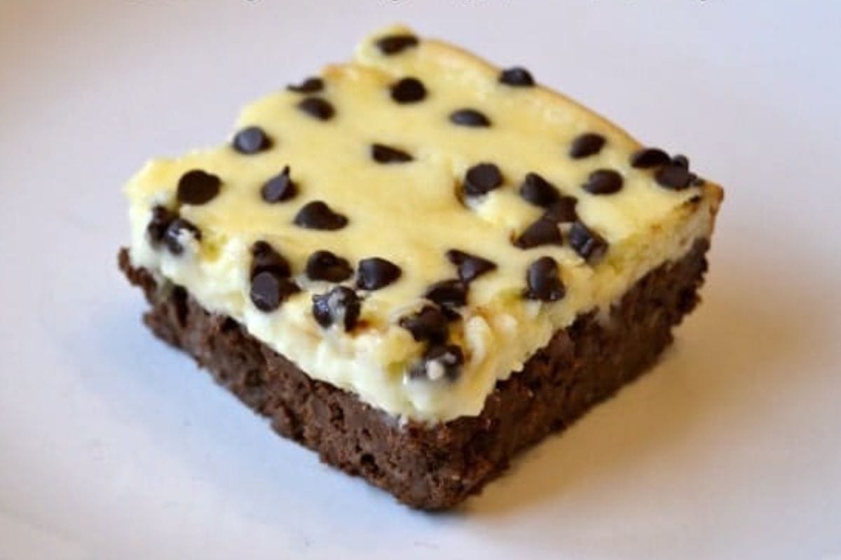 A square piece of guilt-free cheesecake brownies with a brownie base, a creamy cheesecake layer, and chocolate chips sprinkled on top, placed on a white surface.