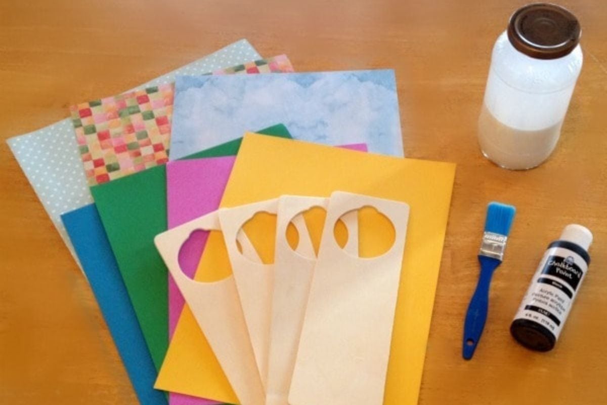 Assorted colored and patterned paper, blank wooden DIY door hanger, a paintbrush, a bottle of paint, and a jar of liquid are arranged on a wooden surface.