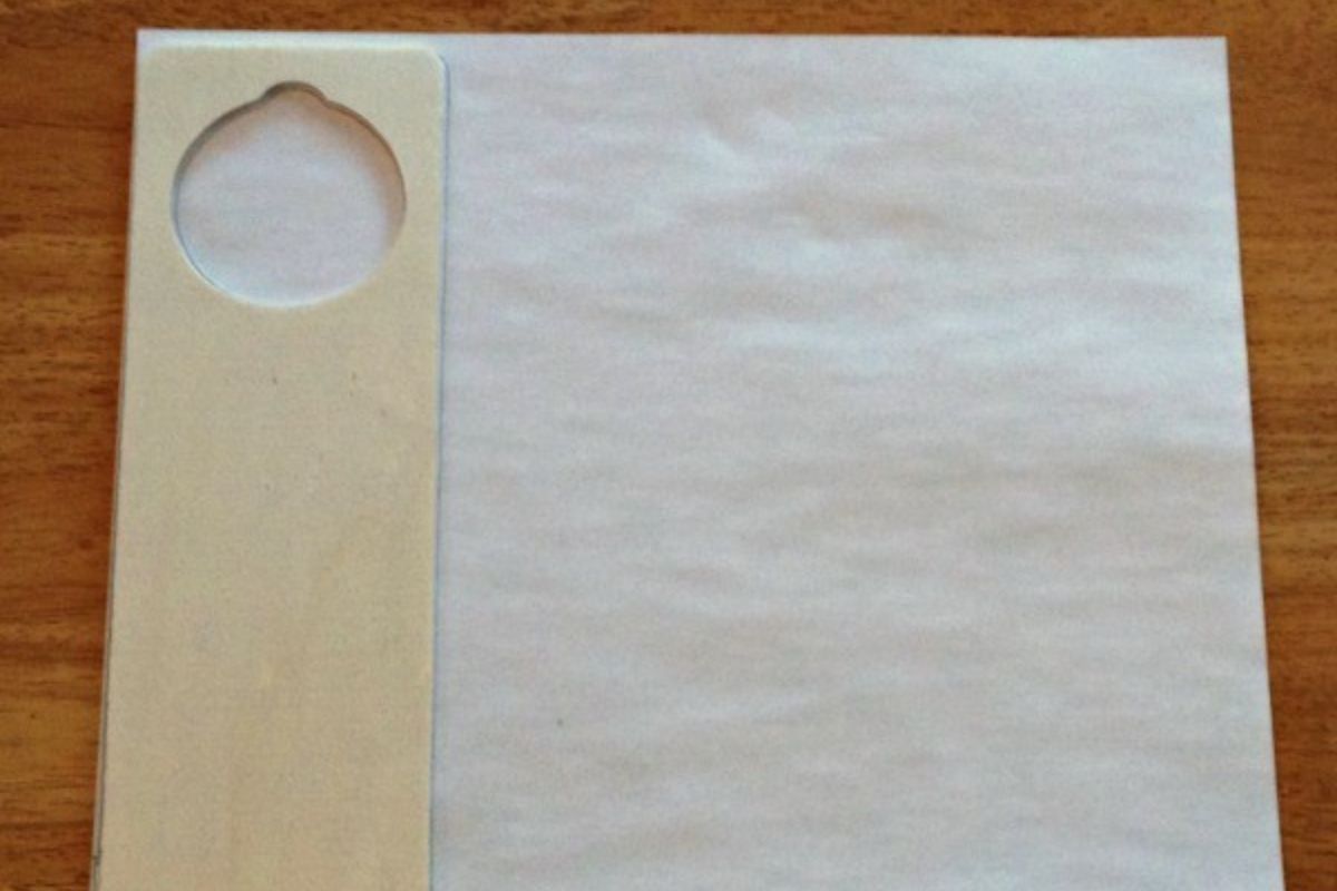 A square napkin with a wide border on the left featuring a circular cutout near the top, reminiscent of a DIY door hanger, placed on a wooden surface.