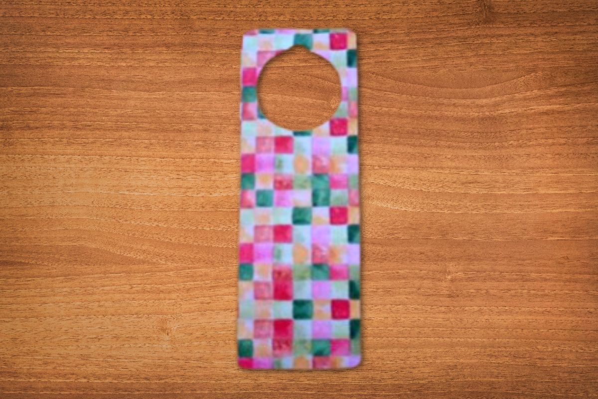 A DIY door hanger, rectangular with a circular hole at the top and a colorful checkered pattern, is placed on a wooden surface.