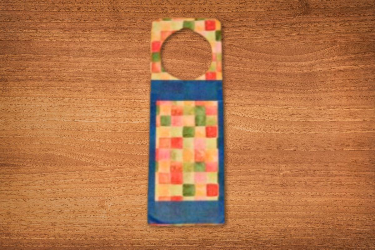 A DIY door hanger, rectangular with a circular cutout at the top, features a colorful square pattern bordered by a solid blue section, displayed on a wooden surface.