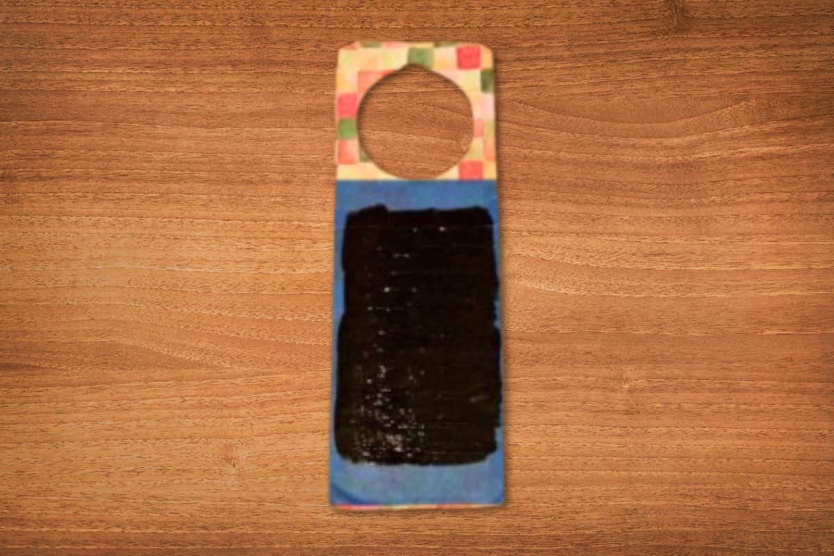 A rectangular wooden DIY door hanger with a circular hole at the top, painted with a colorful border and a large black rectangle in the center, placed on a wooden surface.