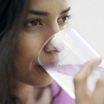 A woman drinks from a clear glass of water, holding the glass near her lips with her face in close-up view, evoking the refreshing simplicity of Zarbee's natural wellness.