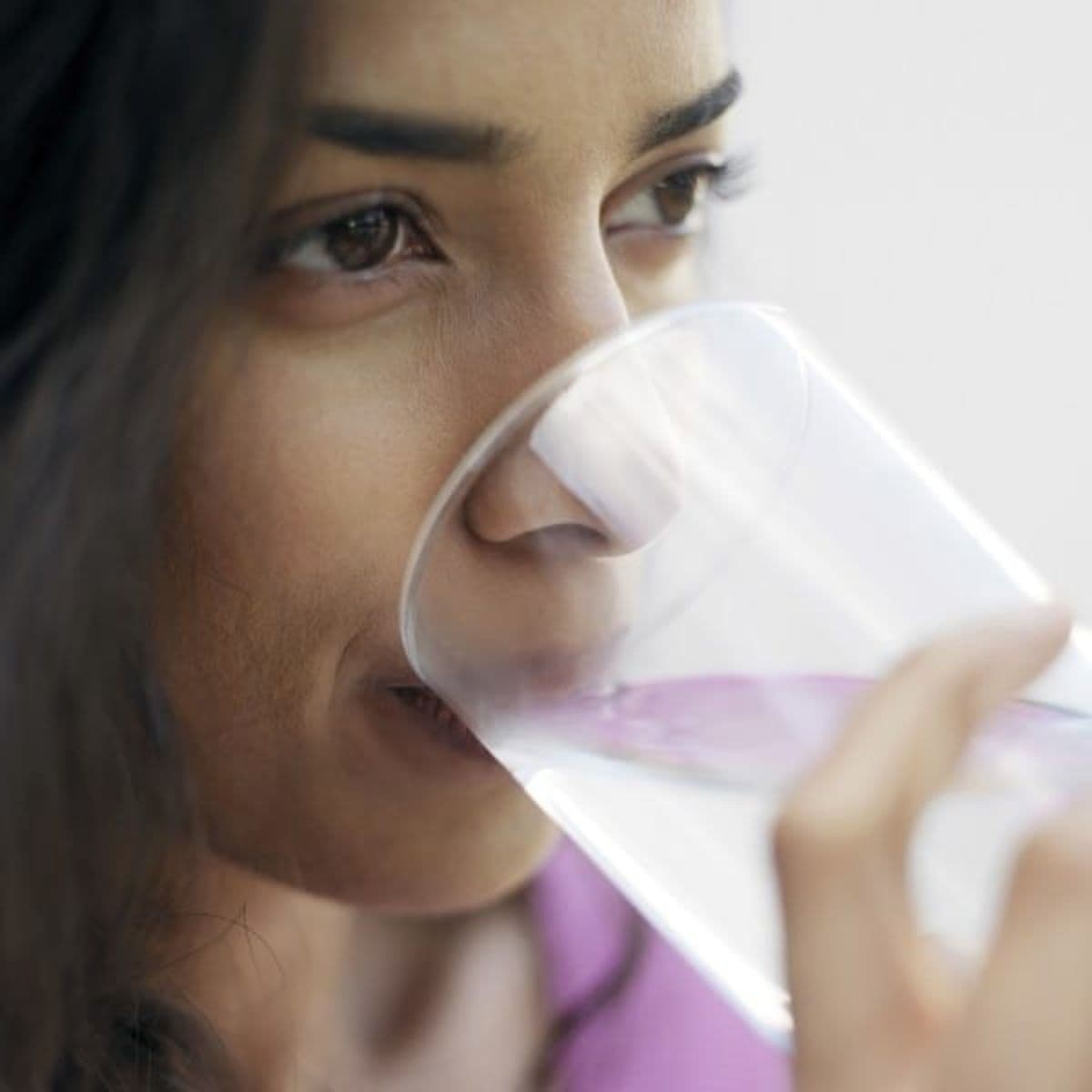 A woman drinks from a clear glass of water, holding the glass near her lips with her face in close-up view, evoking the refreshing simplicity of Zarbee's natural wellness.