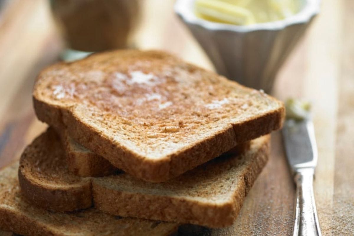 Three slices of Roman Meal bread are stacked on a wooden surface, with a butter knife and a bowl containing butter.