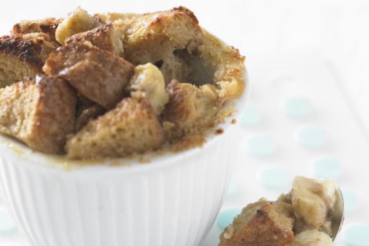 A close-up of bread pudding, featuring chunky pieces in a white ramekin and a spoonful of pudding resting beside it.