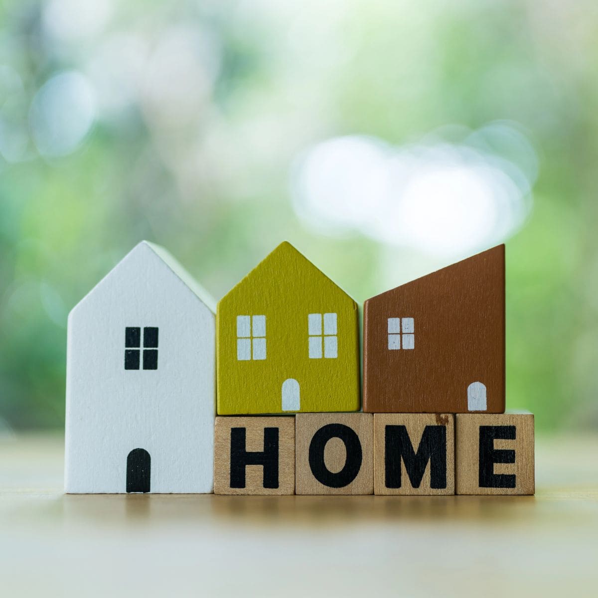 Three small wooden house models stand behind four wooden blocks spelling "HOME" in black letters, symbolizing the journey of buying a home, set against a blurred green background.