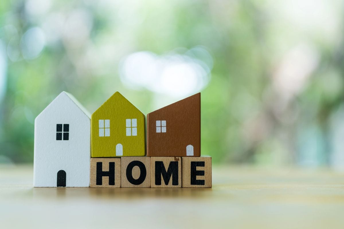 Three small wooden house models in white, yellow, and brown stand behind wooden blocks spelling "HOME".