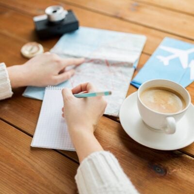 Person planning a trip with a map, camera, airplane tickets, notepad featuring tips for foreign travel, and a cup of coffee on a wooden table.