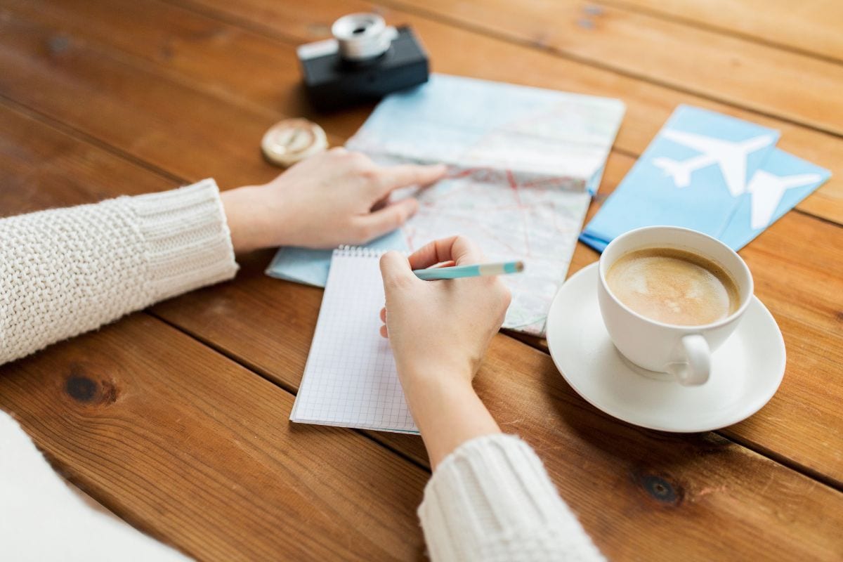 Person planning a trip with a map, jotting down tips for foreign travel in a notepad, with a cup of coffee, camera, and airplane tickets neatly arranged on a wooden table.