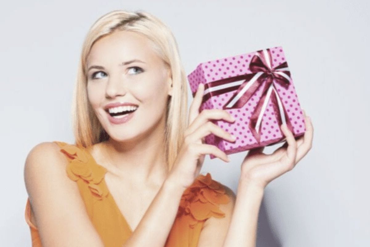 A woman with blonde hair smiles and holds a pink gift box with a bow, looking to the side against a plain background.