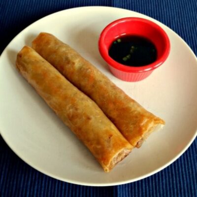 Two crispy lumpia spring rolls on a white plate with a small red cup of dipping sauce, set on a dark blue surface.