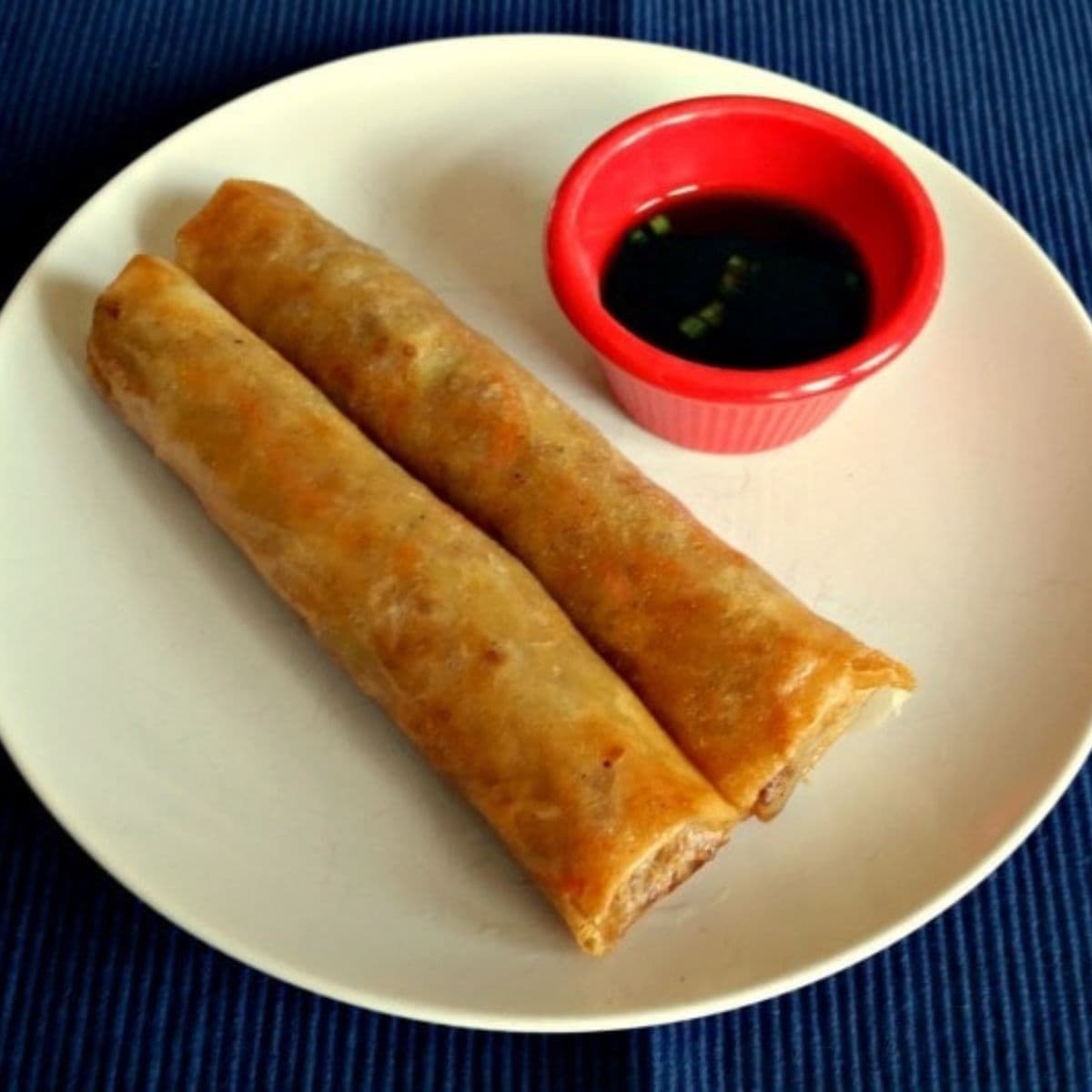 Two crispy lumpia spring rolls on a white plate with a small red cup of dipping sauce, set on a dark blue surface.