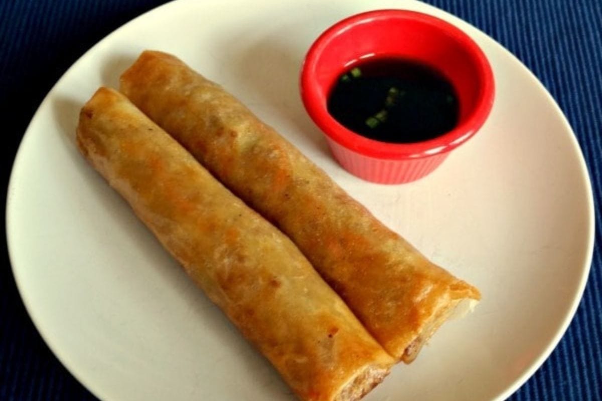 Two crispy lumpia spring rolls on a white plate, served with a small red bowl of dipping sauce.
