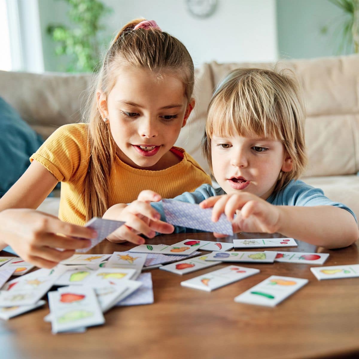 Two children sit at a table playing an educational game, matching illustrated cards and focusing intently on the game pieces in their hands.