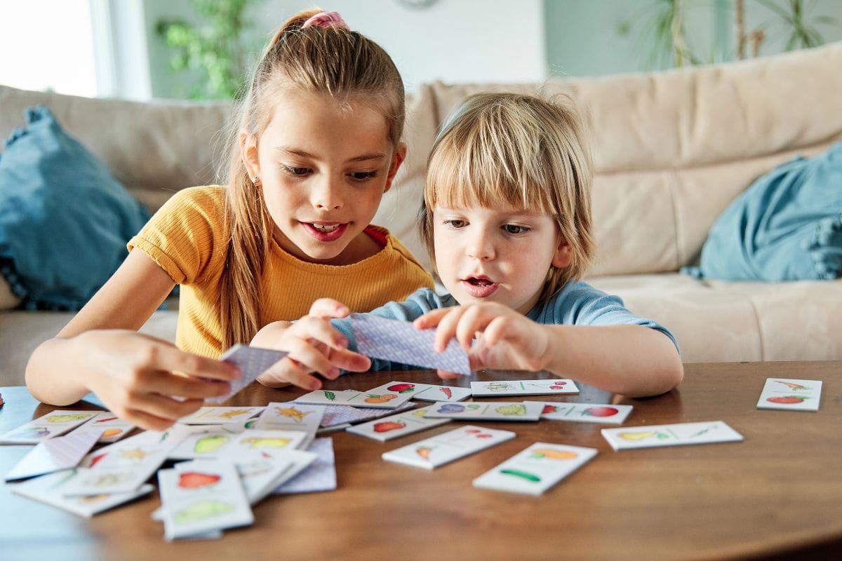 Two children sit at a table in the living room, playing an educational game with illustrated cards featuring various objects and images.