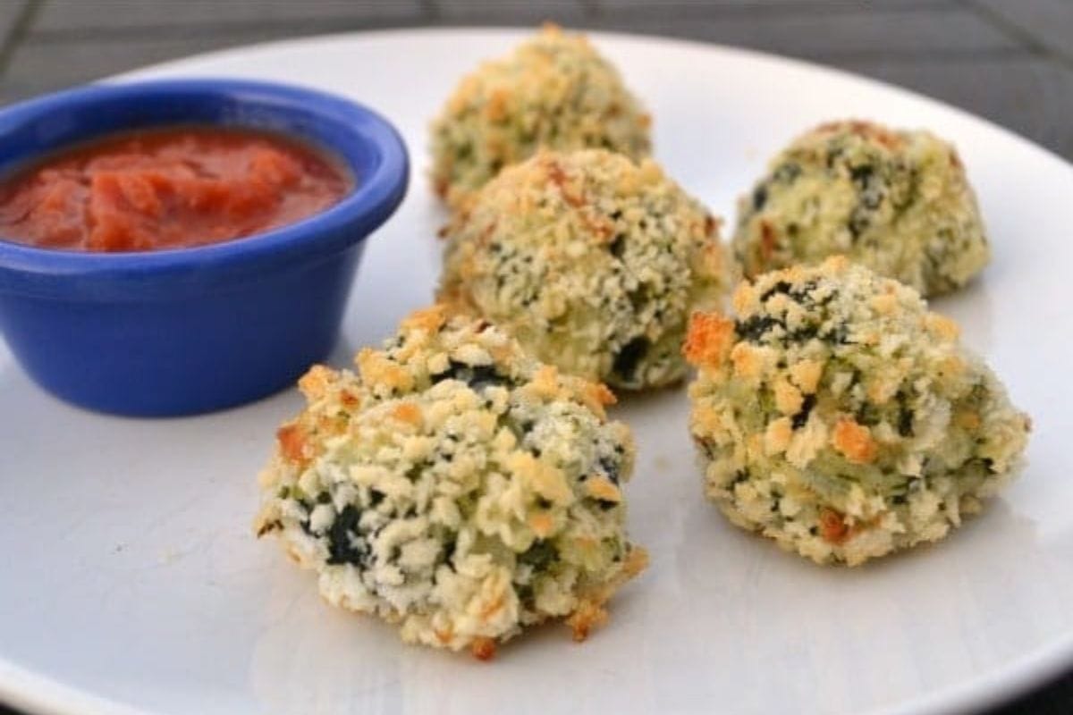 Four breaded, baked spinach rice appetizer balls on a white plate with a small blue cup of red dipping sauce.