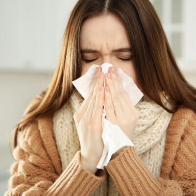 A woman in a brown sweater and scarf holds a tissue to her nose, appearing to sneeze or blow her nose indoors—an all too familiar scene during allergy season.