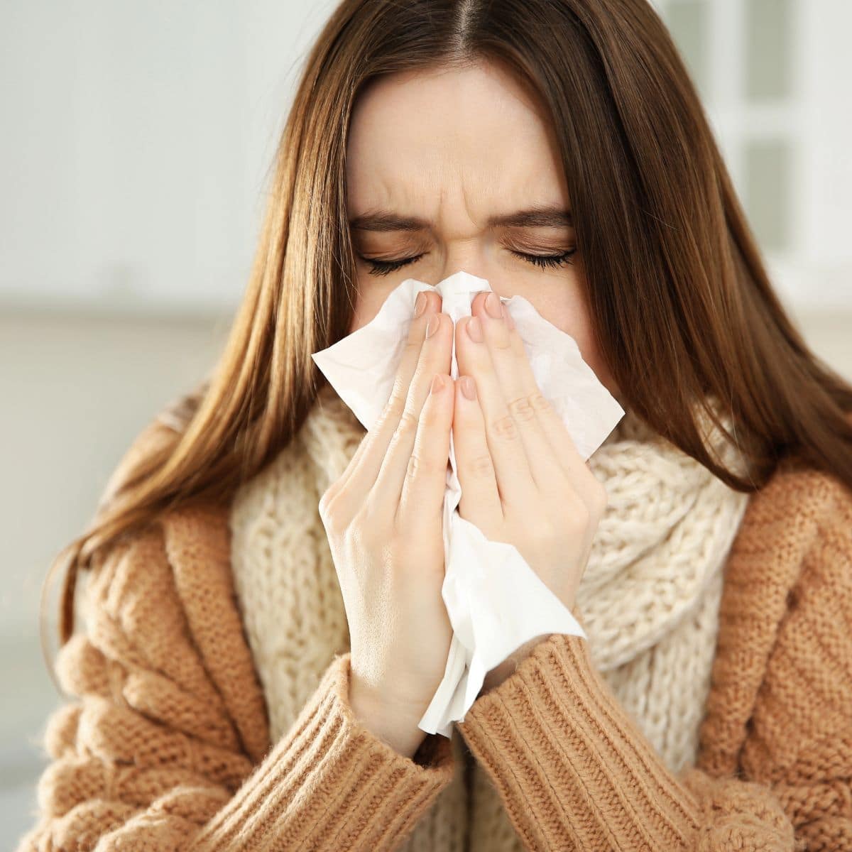 A woman in a brown sweater and scarf holds a tissue to her nose, appearing to sneeze or blow her nose indoors—an all too familiar scene during allergy season.
