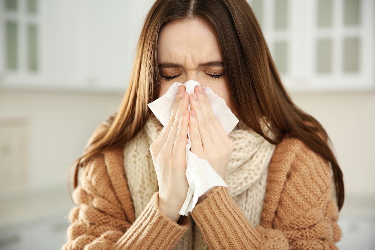 A woman wearing a sweater and scarf is indoors, holding a tissue to her nose and appearing to sneeze.