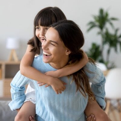 Smiling woman giving a piggyback ride to a laughing young girl in a bright, modern living room.