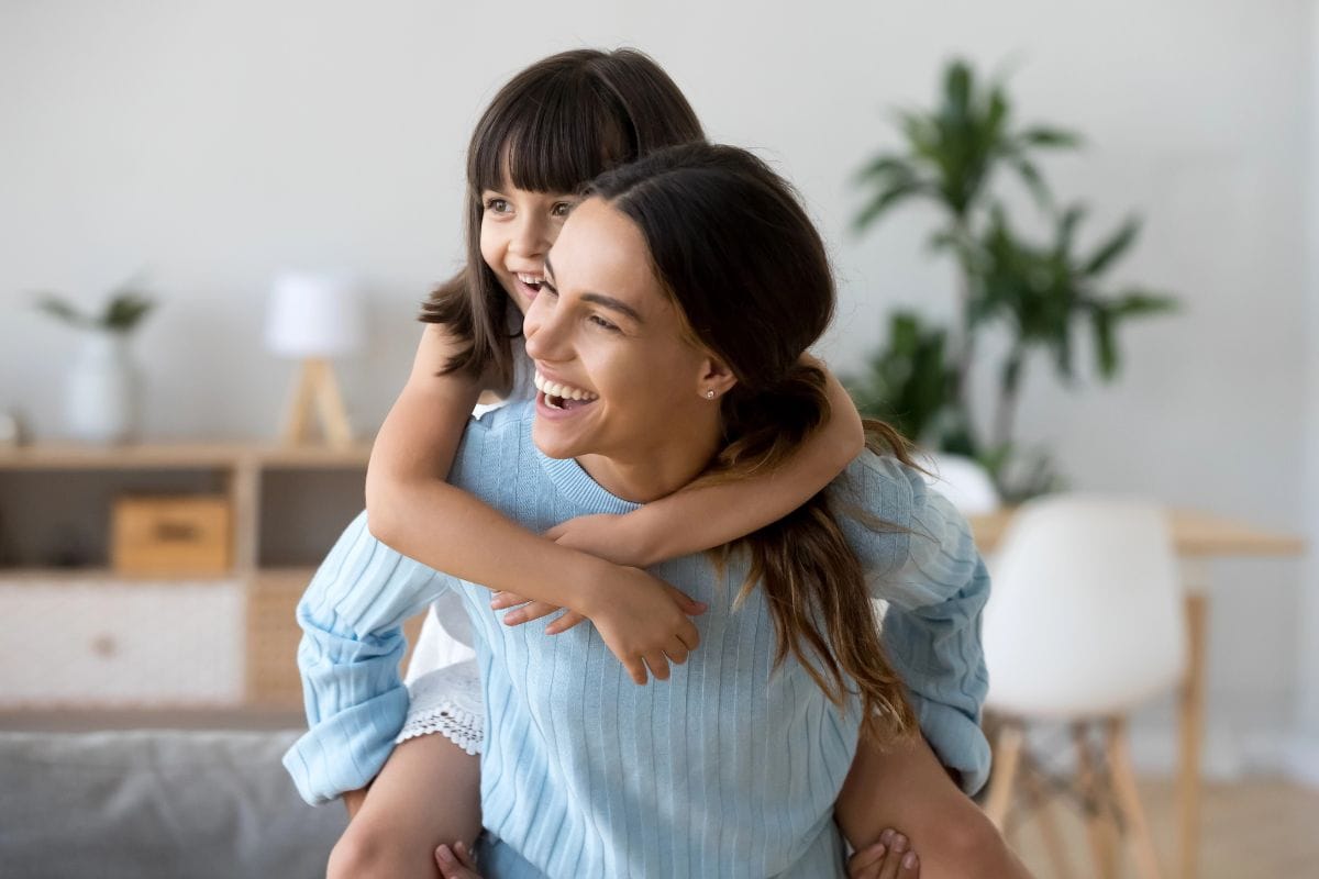 A young woman smiling while giving a piggyback ride to a laughing girl in a bright, modern living room.