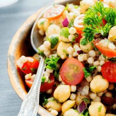 A bowl of couscous salad with cherry tomatoes, chickpeas, red onion, parsley, and a spoon—perfect for meal prep or carb cycling routines.