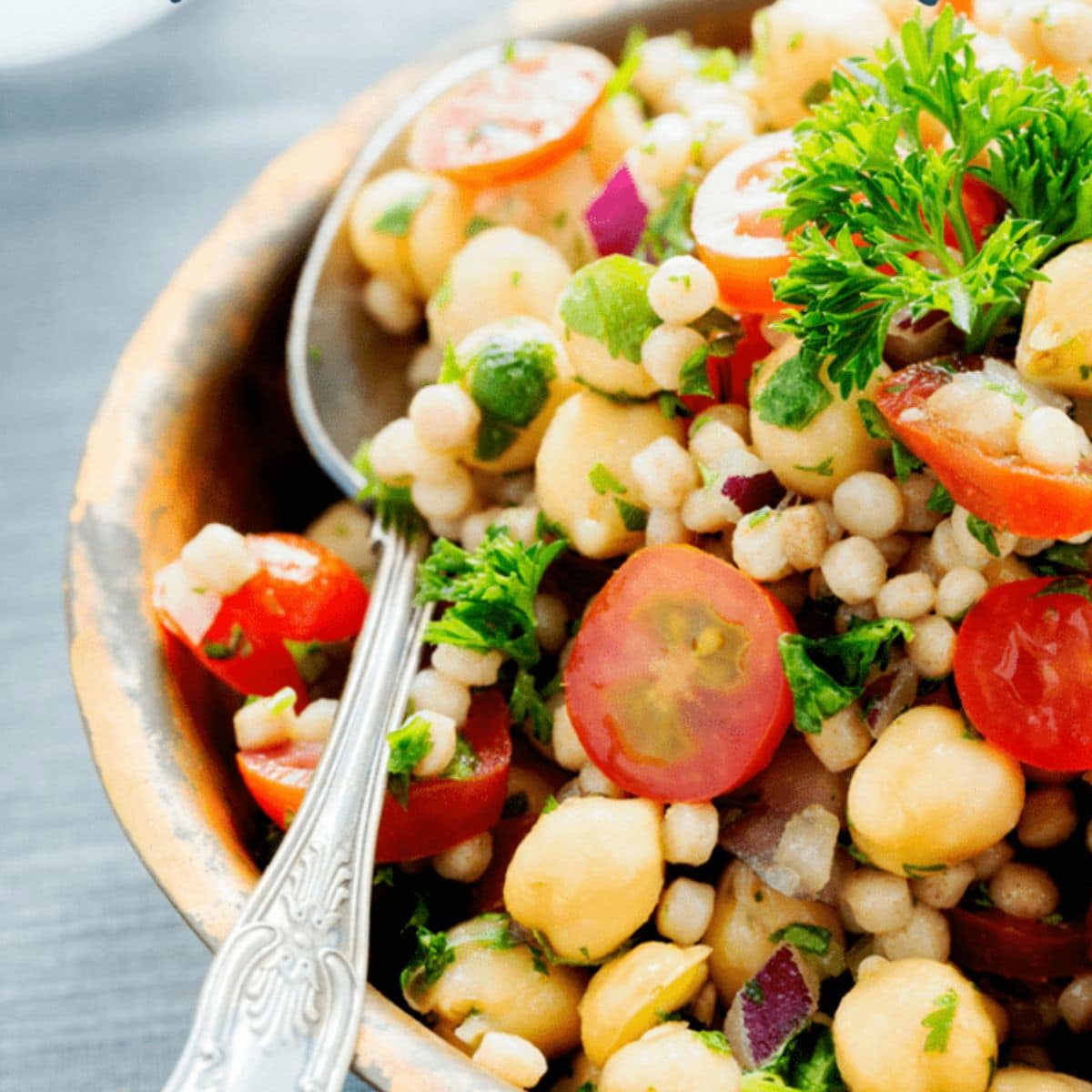 A bowl of couscous salad with cherry tomatoes, chickpeas, red onion, parsley, and a spoon—perfect for meal prep or carb cycling routines.
