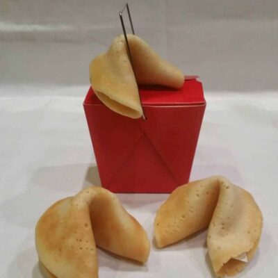 Three homemade fortune cookies, one resting on a closed red takeout box and two placed on a white surface in front of it.