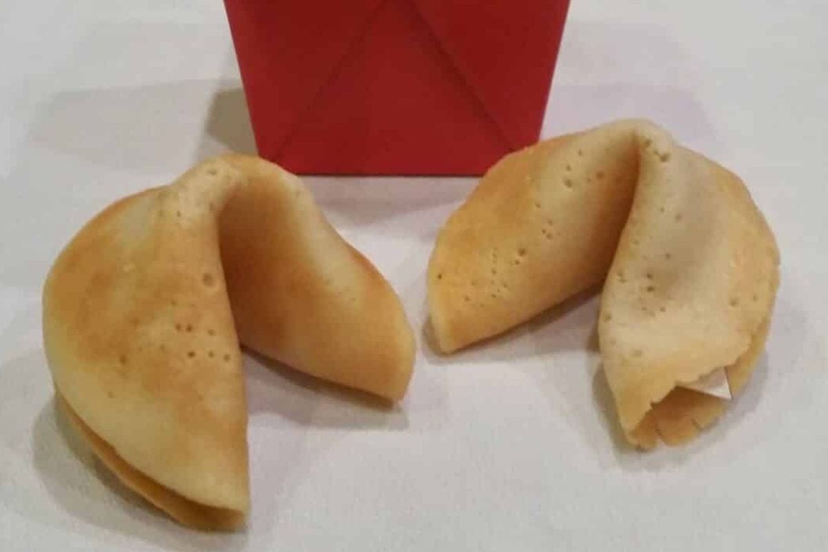 Two homemade fortune cookies are placed on a white surface with a red takeout box in the background.
