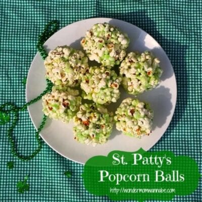 A white plate with seven St. Patty’s popcorn balls sits on a green checkered cloth, surrounded by green beads and a shamrock decoration. Text reads “St. Patty’s Popcorn Balls.”.