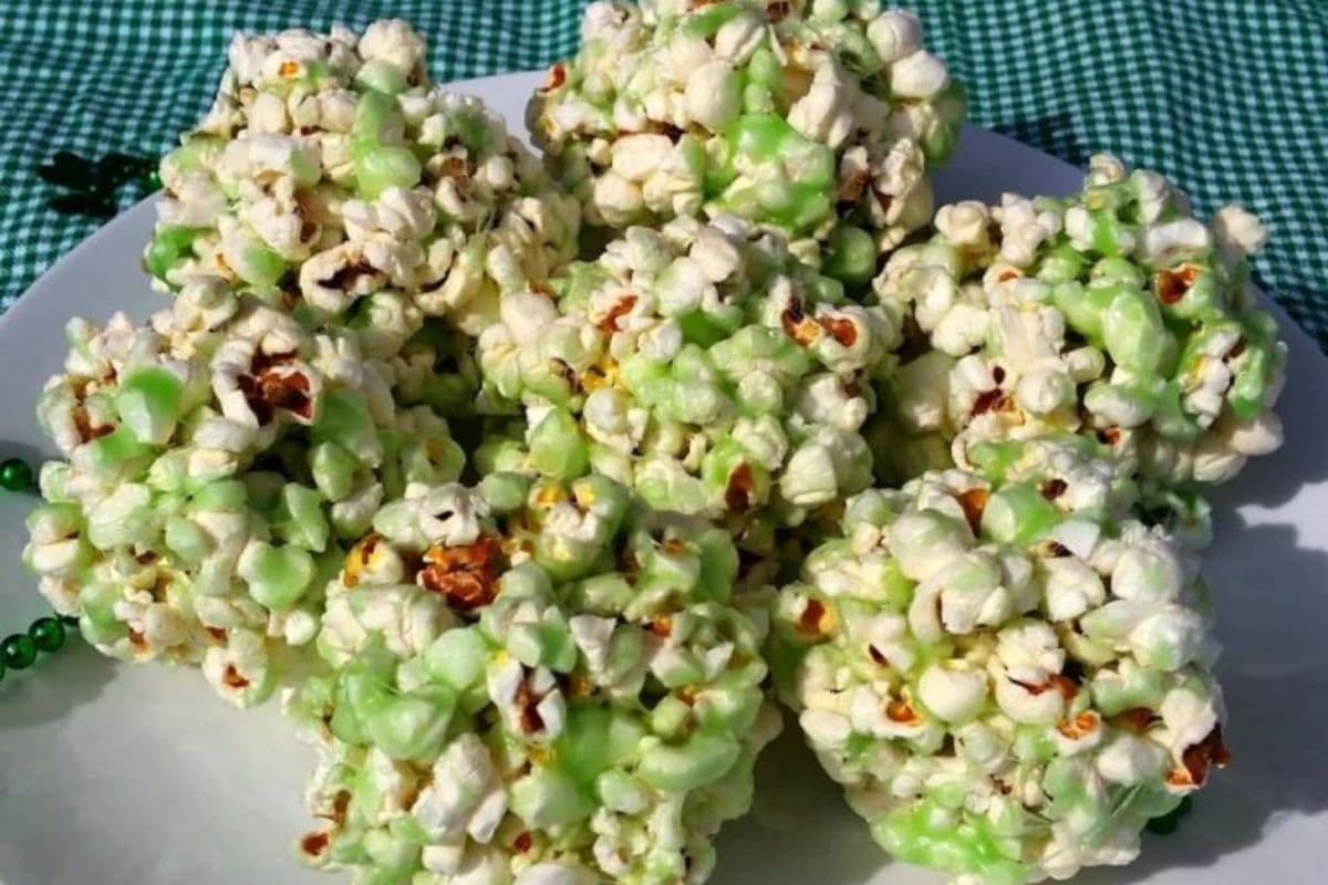 A white plate holds several St. Patty's popcorn balls, their green-tinted coating echoing the festive spirit, set on a green checkered cloth background.
